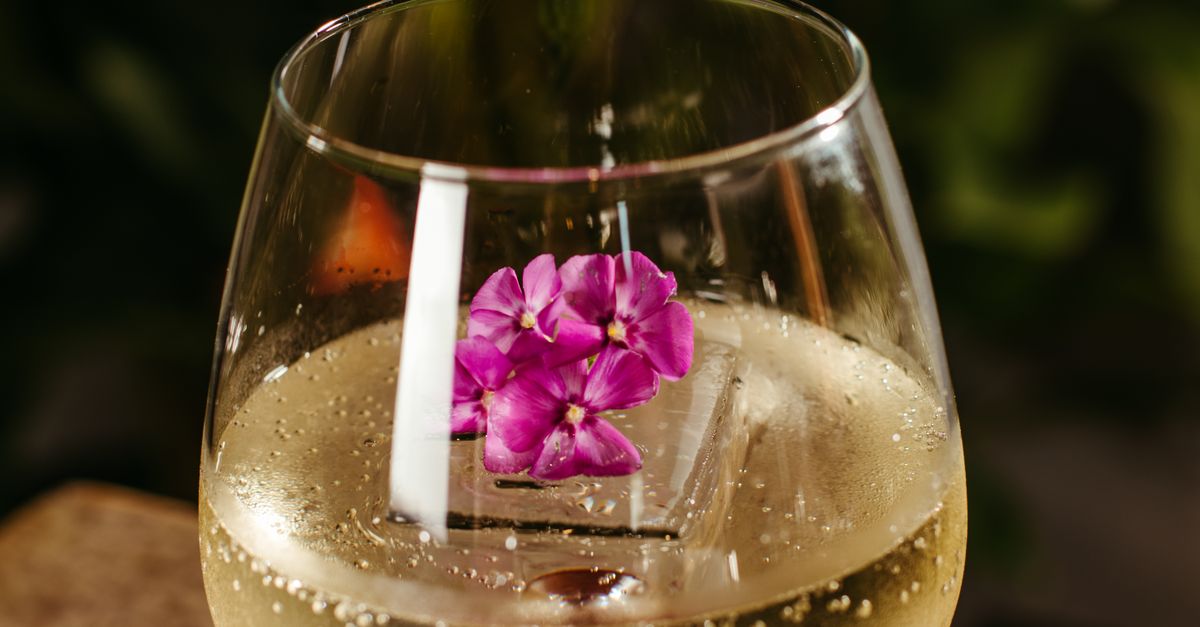 Close-up of a bubbly white wine in a glass with a purple flower garnish.