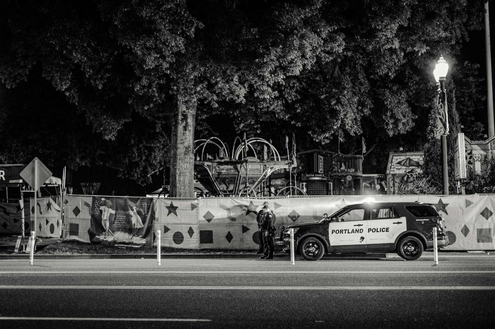 Greyscale Photo of Two Policemen Standing Infront of a Parked Police Car