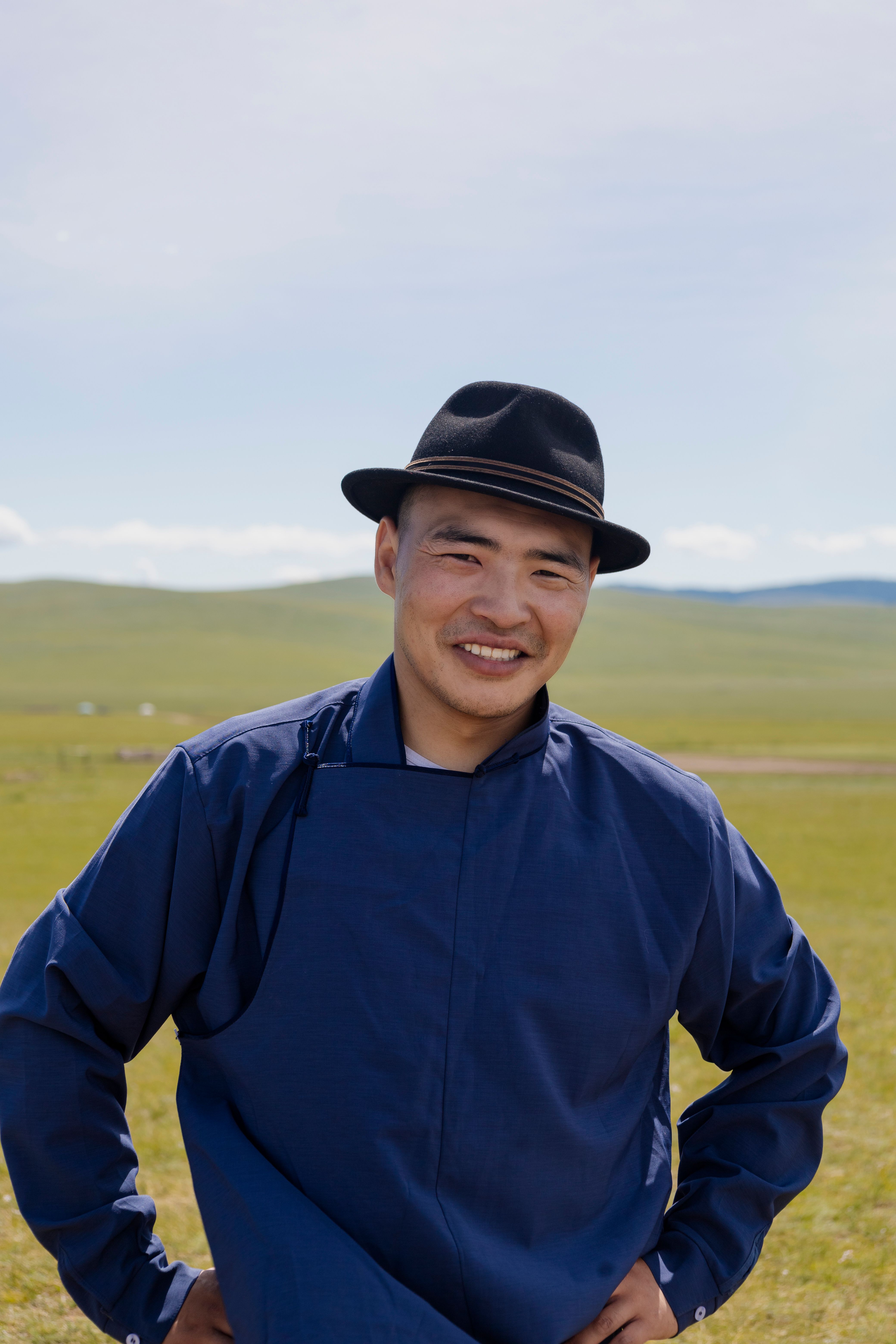 Smiling man in traditional blue attire stands confidently in a sunny outdoor setting.