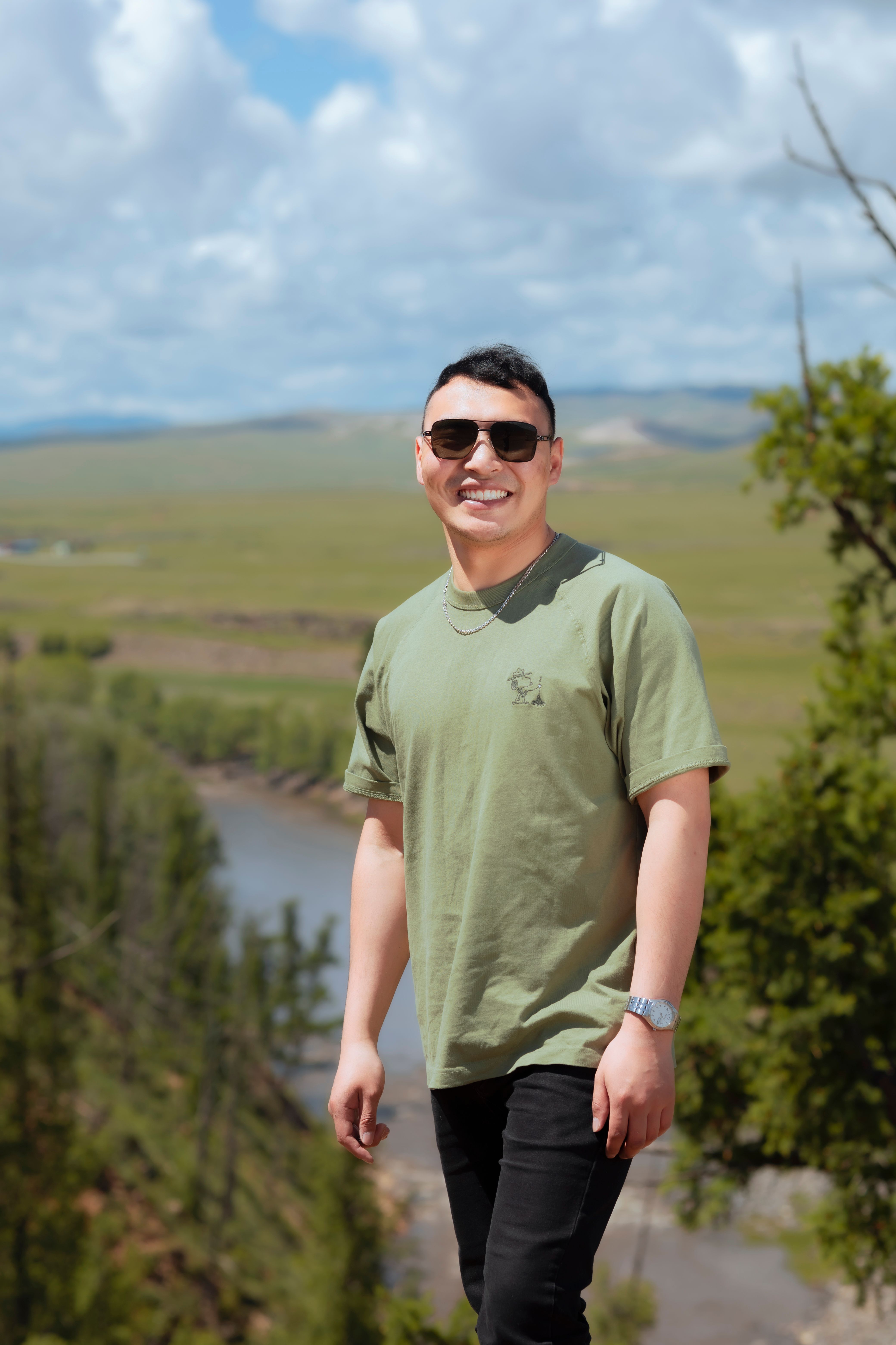 A young man with sunglasses smiling in a lush outdoor landscape.
