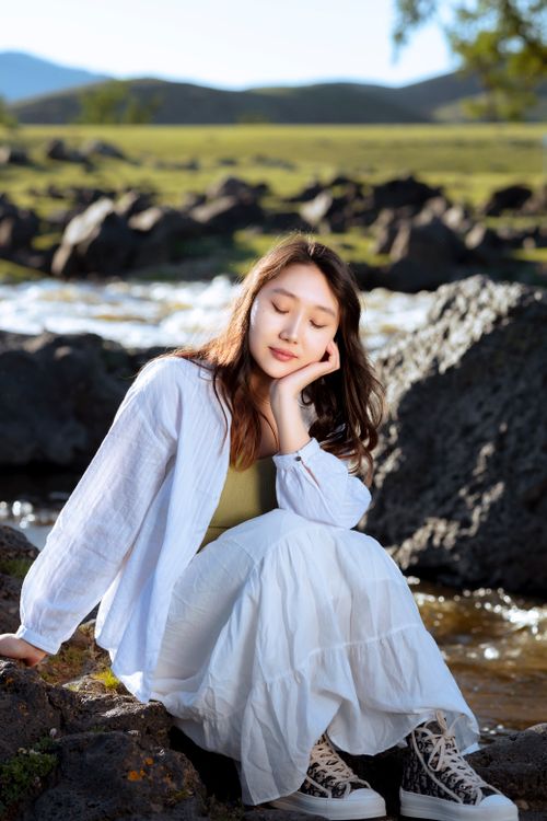 Free A young woman in white clothing resting by a tranquil river with a scenic mountain backdrop. Stock Photo