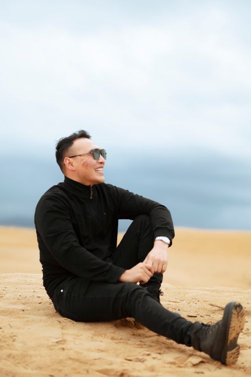 Free Smiling man wearing sunglasses sitting on sandy beach in casual black attire. Stock Photo