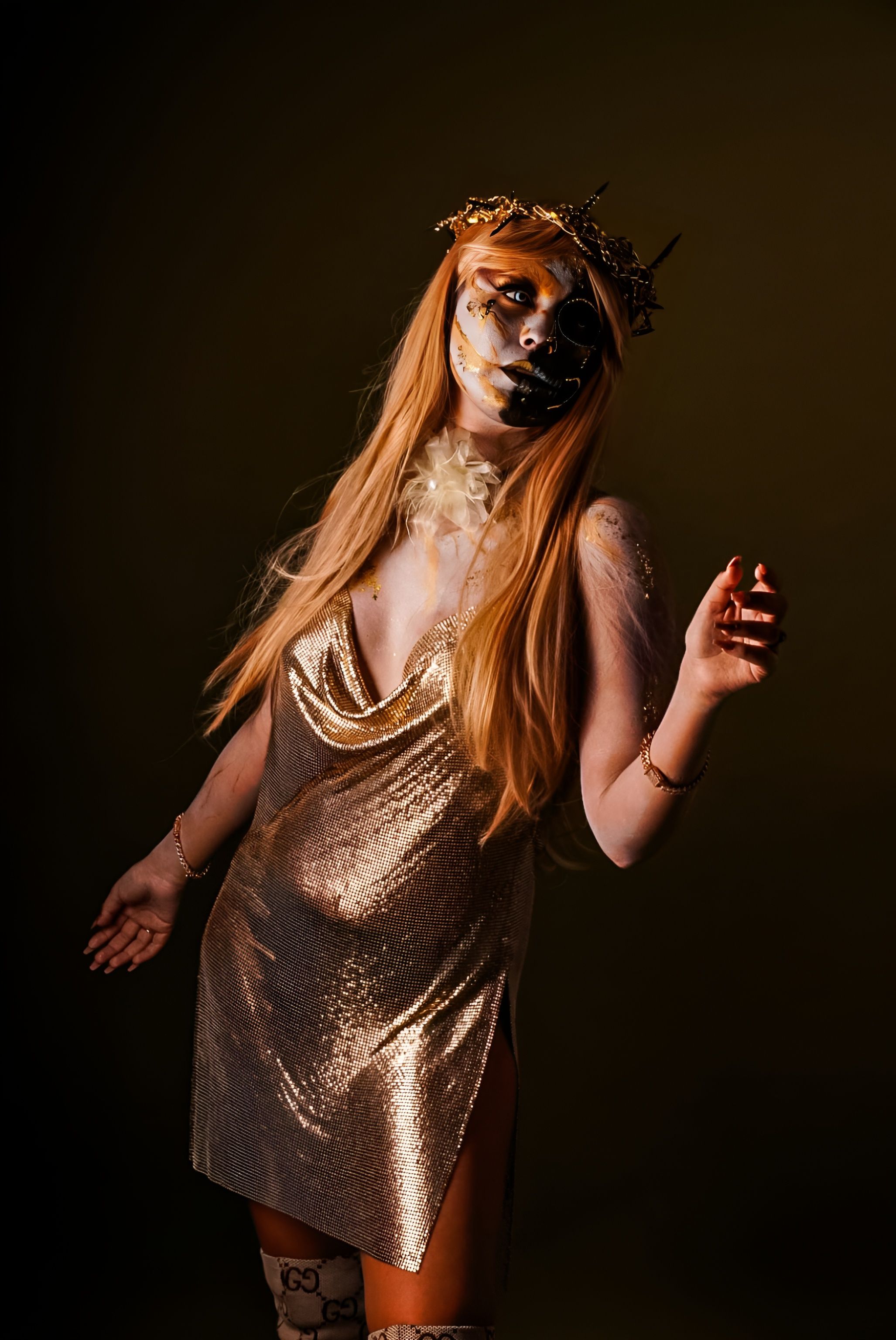 A woman in elaborate artistic makeup and gold dress poses confidently.