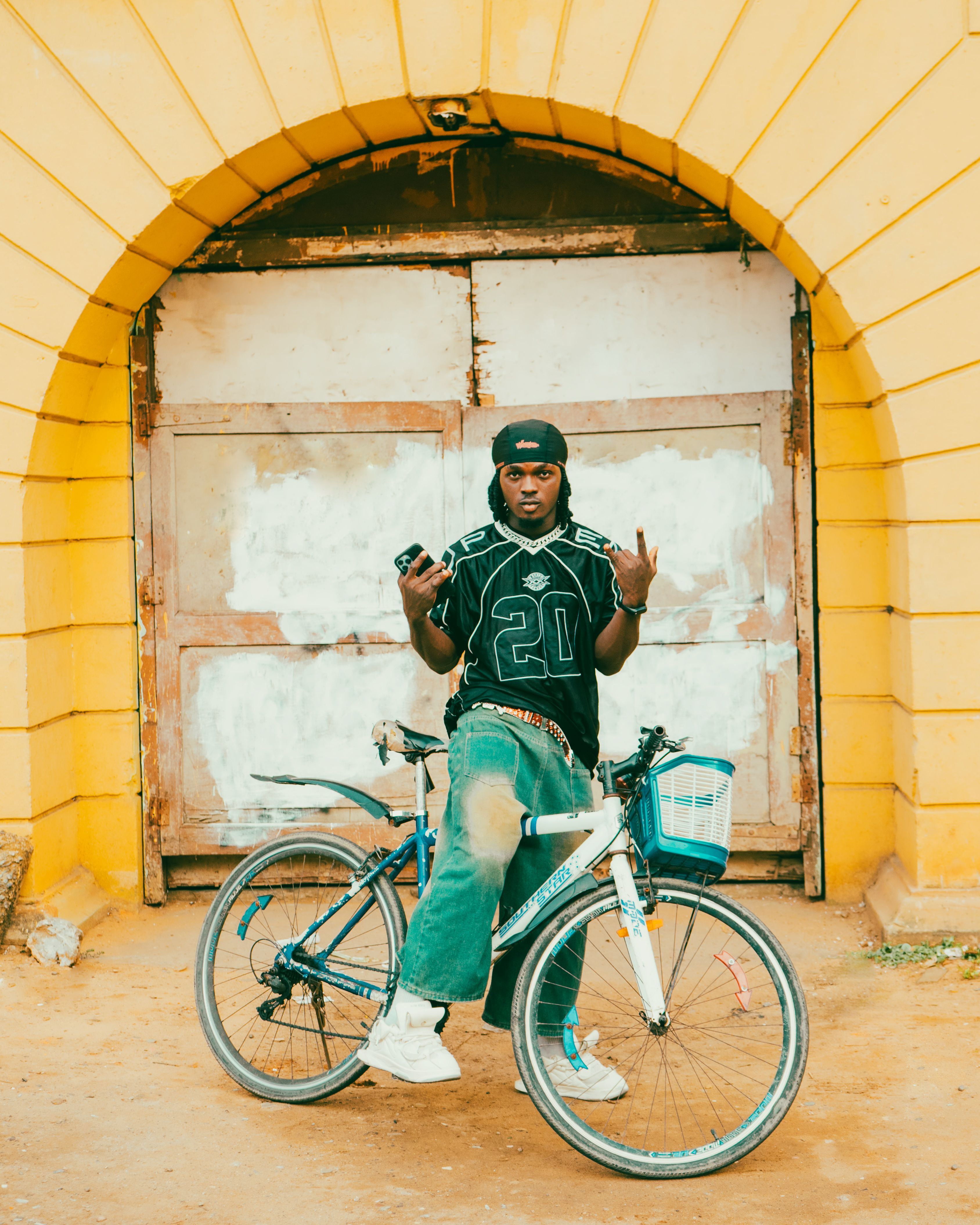 Trendy young adult posing with a bicycle in front of a yellow arched wall.
