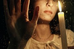 A woman holds a candle behind a rain-spattered window, creating a moody and reflective ambiance.