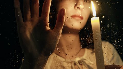 A woman holds a candle behind a rain-spattered window, creating a moody and reflective ambiance.