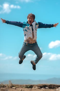 A cheerful young man jumps mid-air against a bright blue sky, exuding joy and energy.