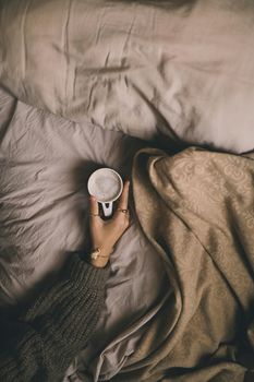Warm cozy morning setting with a hand holding a coffee cup in bed.
