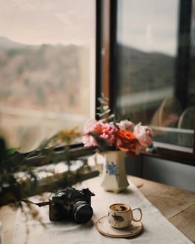 A vintage camera, coffee cup, and flowers on a wooden table by a window.