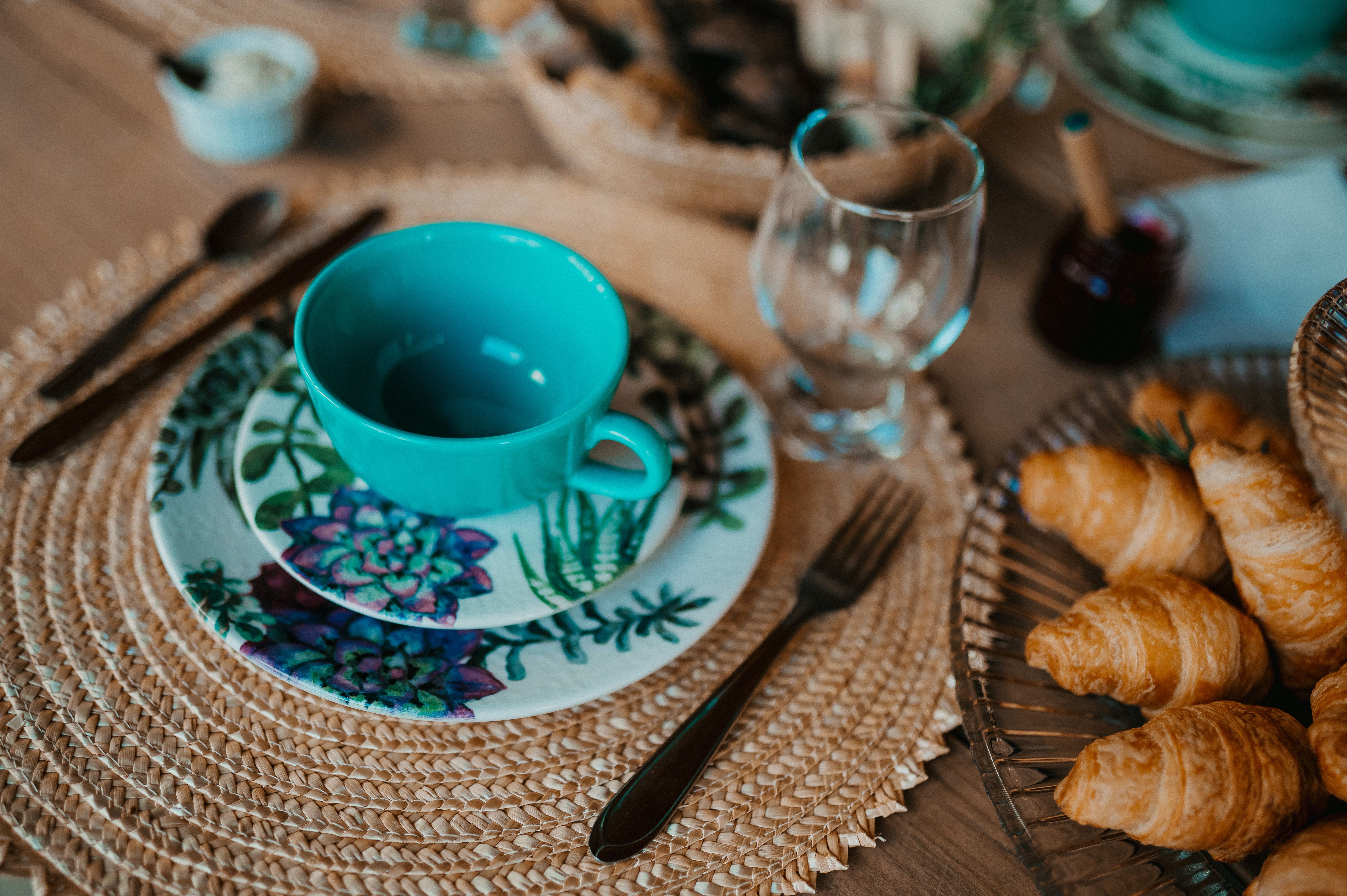 A cozy breakfast setup featuring a teal teacup, plate with floral design, and freshly baked croissants on a woven placemat.