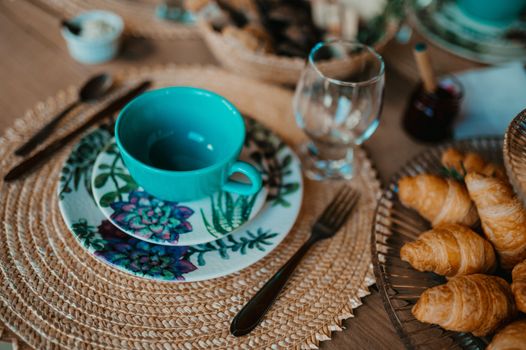 A cozy breakfast setup featuring a teal teacup, plate with floral design, and freshly baked croissants on a woven placemat.