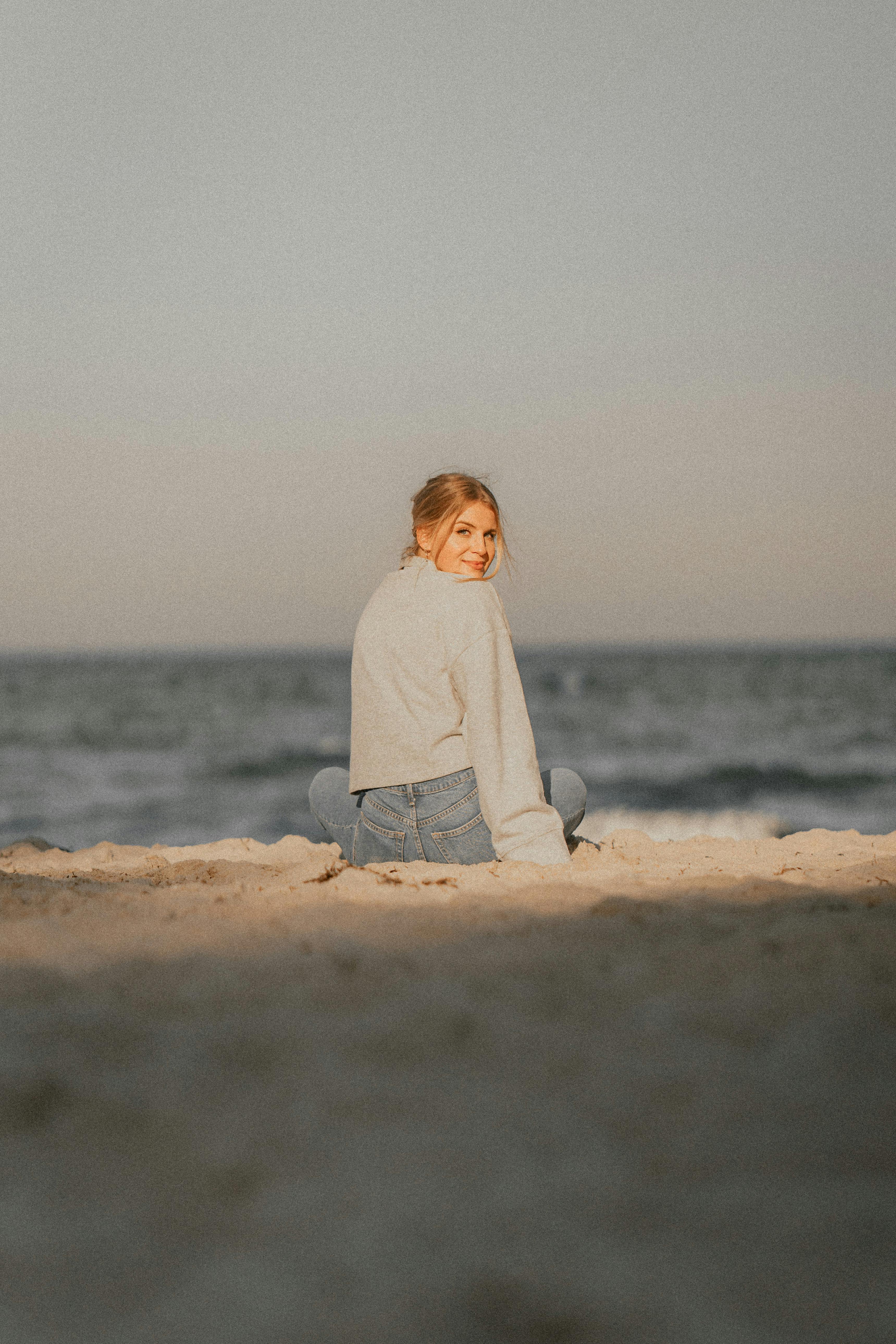 grátis Jovem sorridente sentada na praia ao pôr do sol, de frente para o oceano. Foto profissional