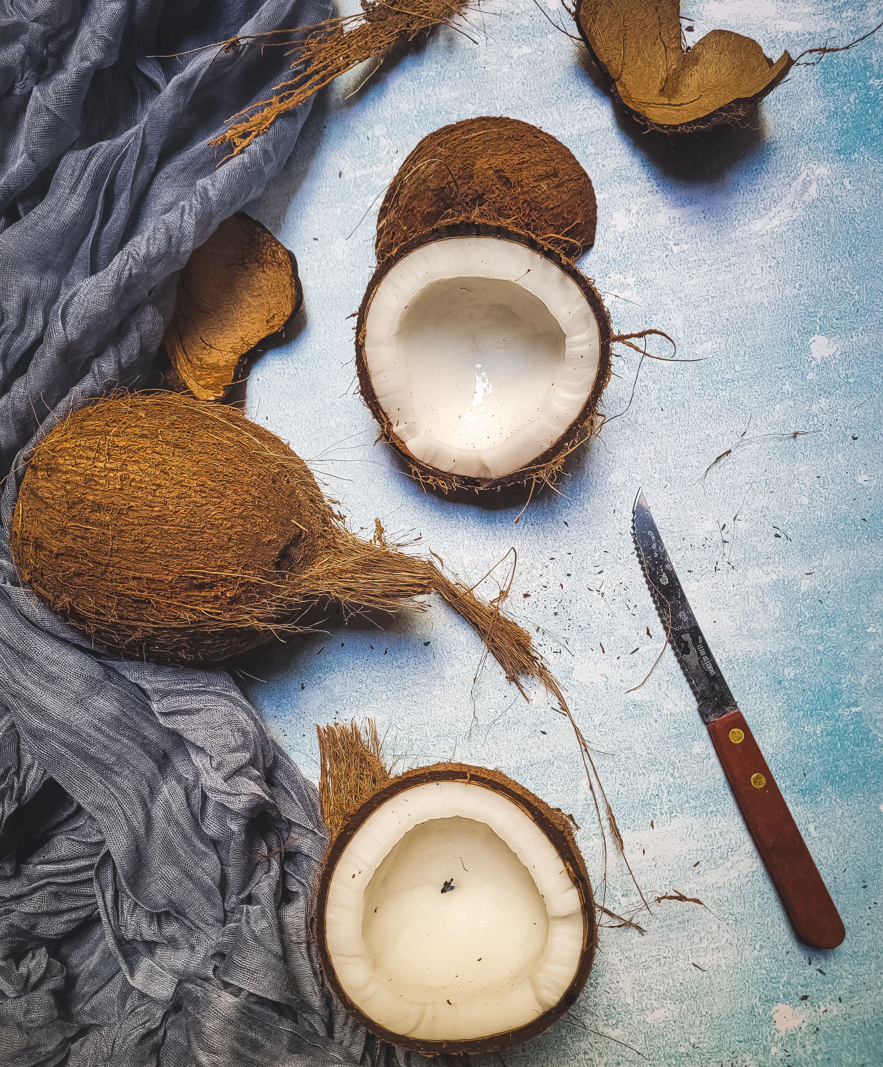 Coconut halves and peeler knife on a rustic surface with fabric draped around for a tropical aesthetic.