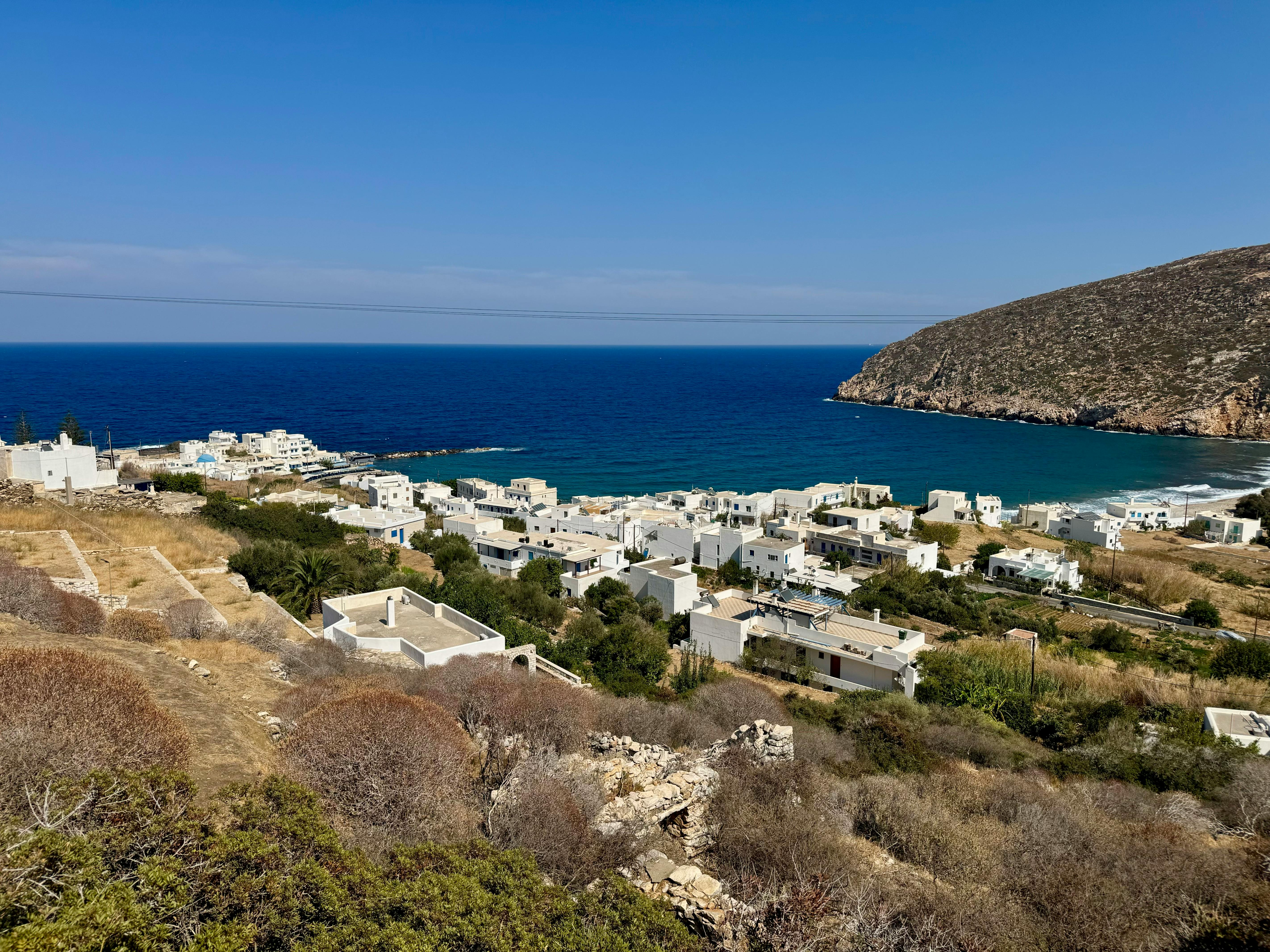 View of a coastal village with white buildings, surrounded by hills and the blue sea under a clear sky.