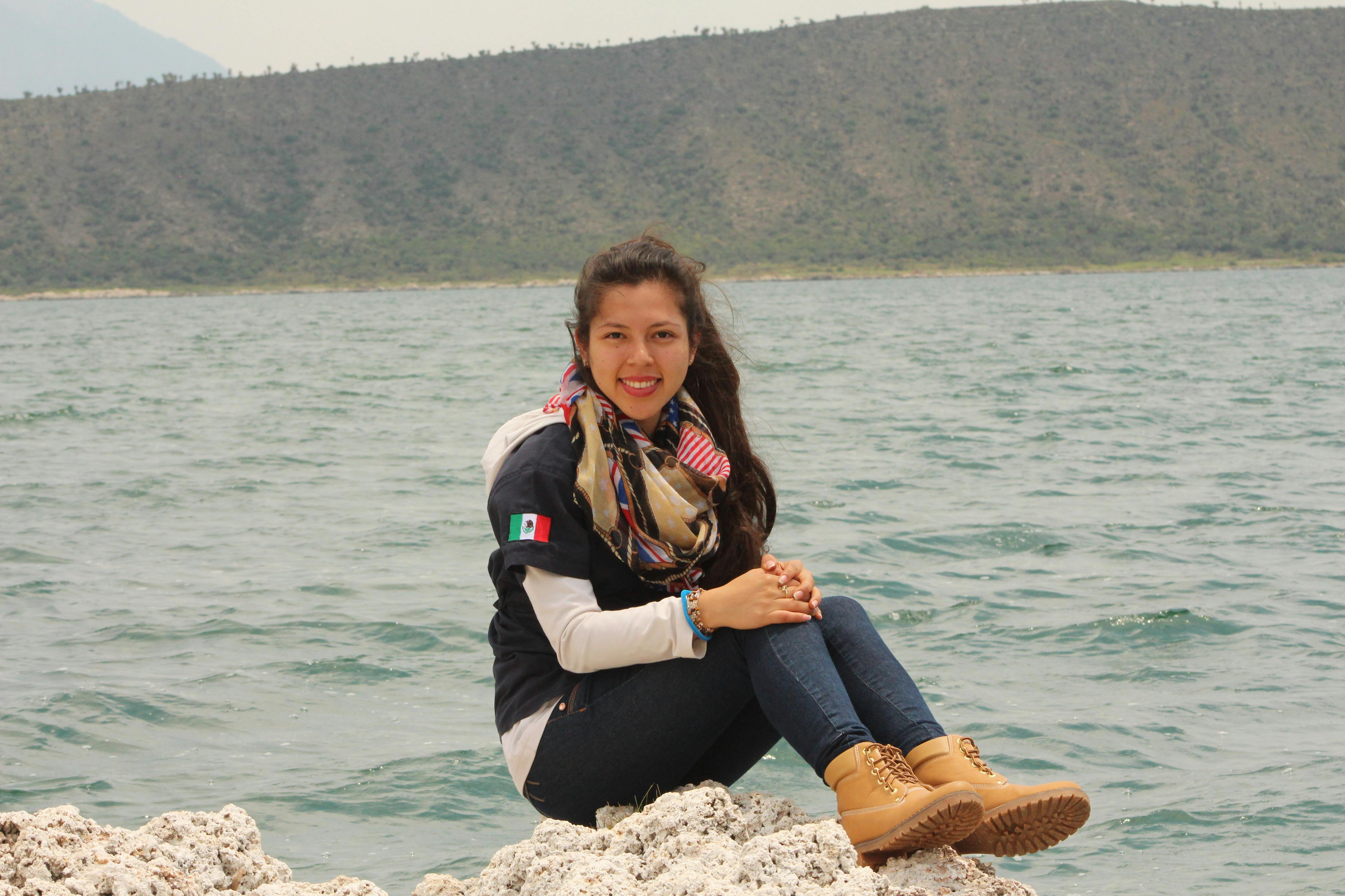 Smiling woman in casual attire sitting by a serene mountain lake on a sunny day.