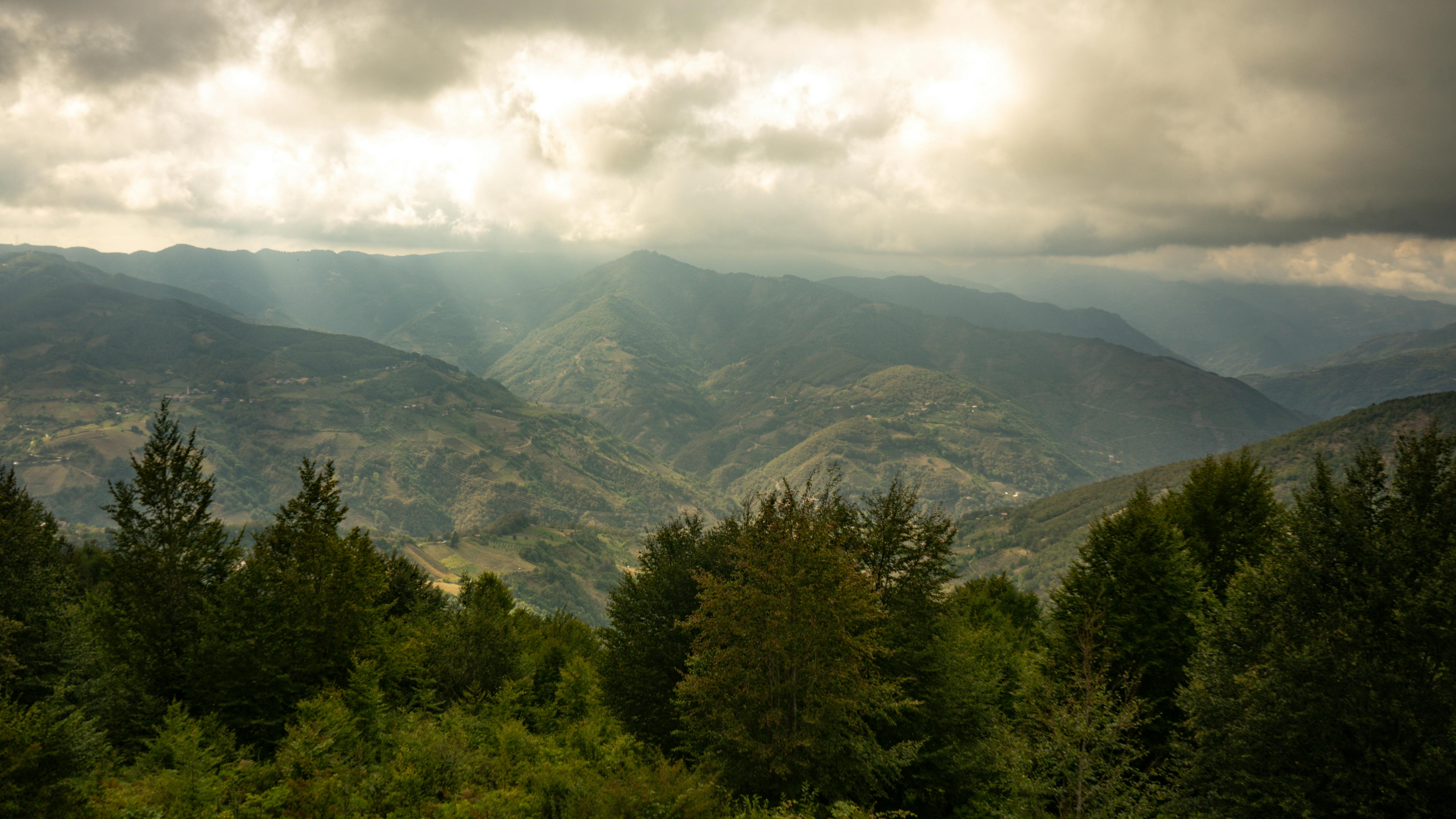Free Beautiful mountainous terrain with lush forests under a dramatic cloudy sky. Stock Photo