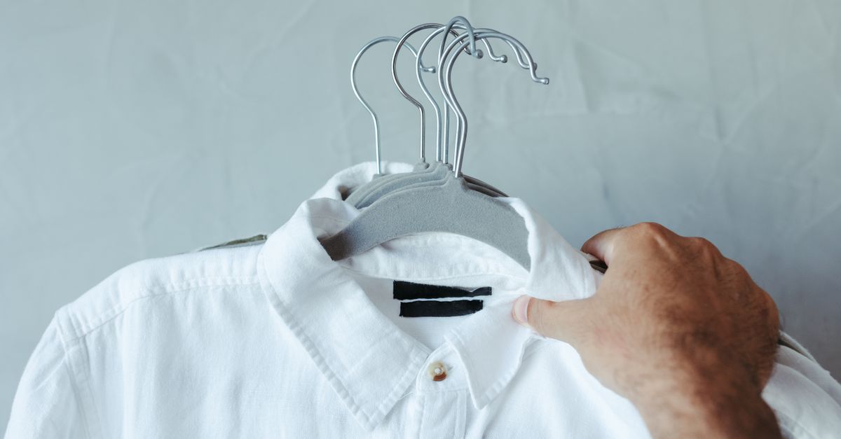 Close-up de uma camisa branca em um cabide segurada por uma mão contra um fundo liso.