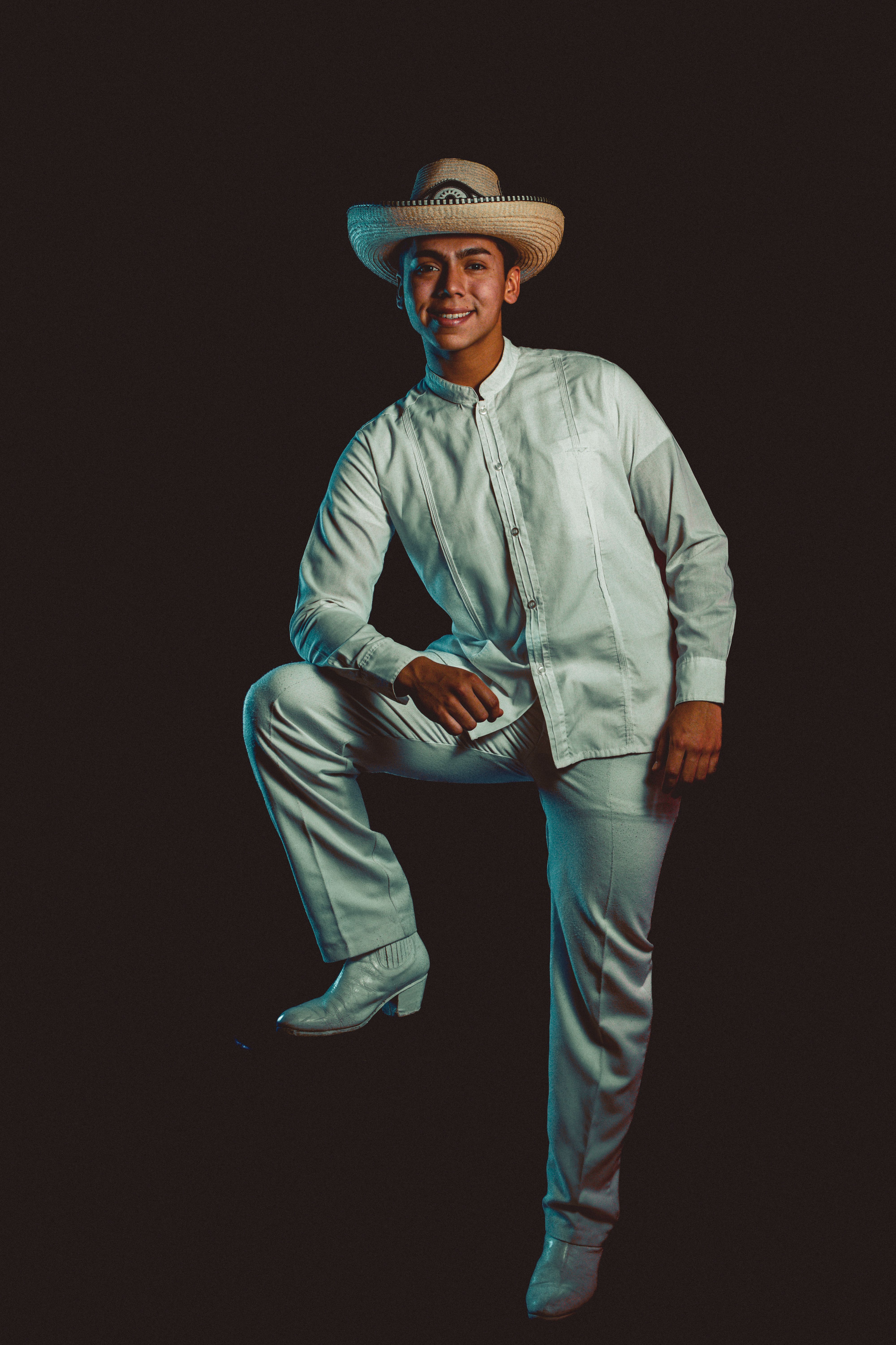 Portrait of a young man wearing traditional white Mexican folk attire and hat, posing against a dark background.