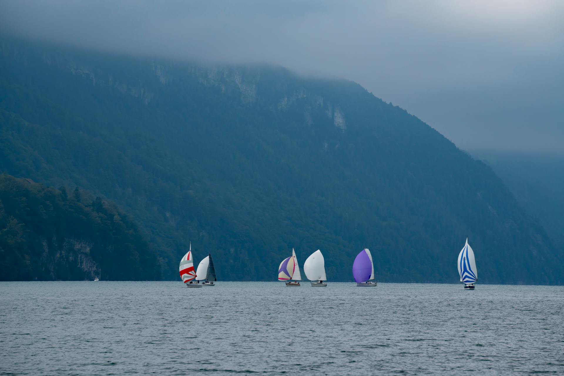 A fleet of sailboats with vibrant sails navigates a calm lake against a mountainous backdrop under a cloudy sky.