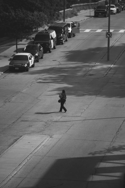 Free A person crossing an urban street in Milwaukee, captured in black and white, with parked cars lining the road. Stock Photo