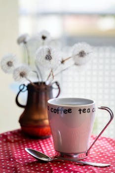 White coffee cup with spoon on polka dot cloth, next to rustic vase with dandelions.