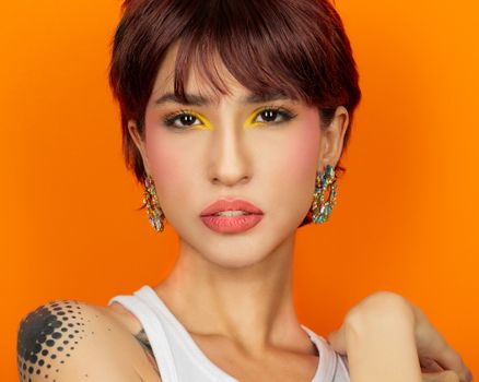 Colorful portrait of a woman with striking makeup and earrings on an orange background.