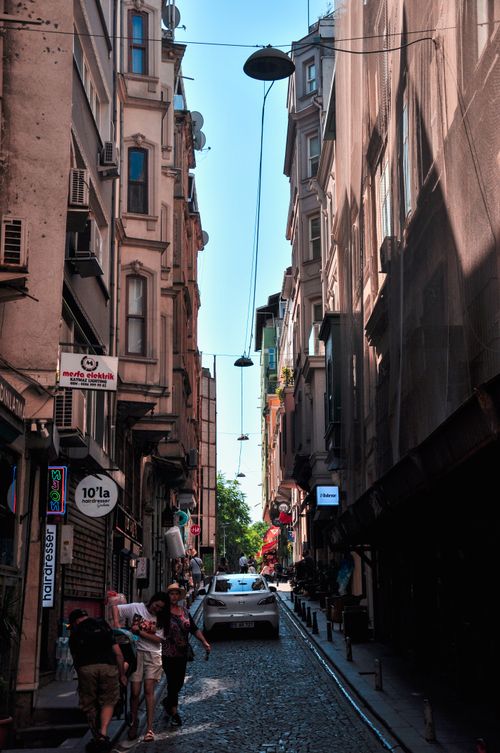 Free A busy narrow street with pedestrians and cars between tall buildings on a sunny day. Stock Photo