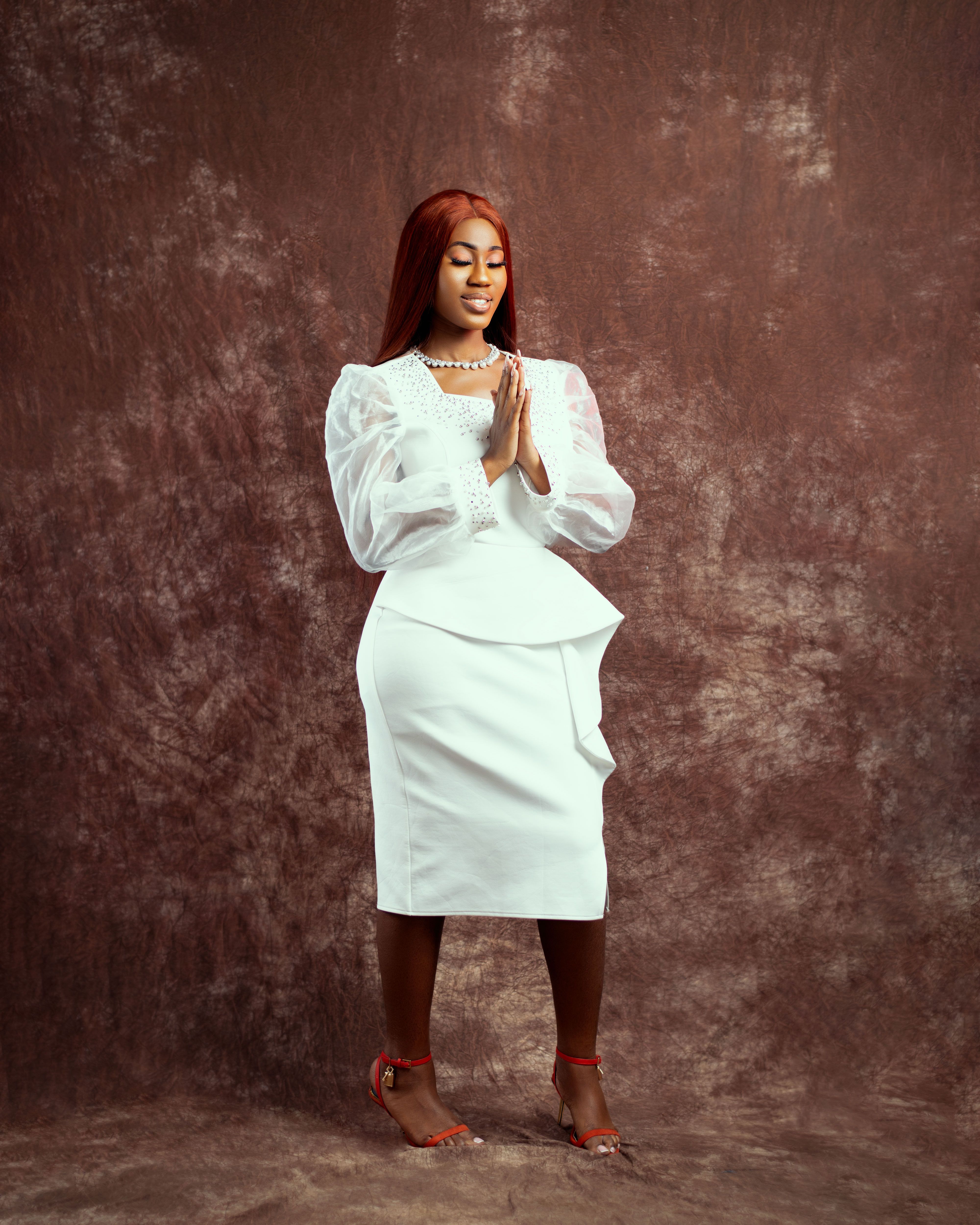 Portrait of a woman in a white outfit with red hair, hands together, against a studio backdrop.