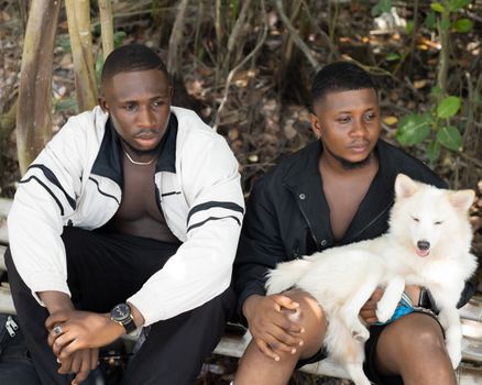 Two men sitting outdoors with a fluffy white dog, enjoying nature.