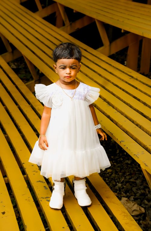 Free A young child stands on yellow wooden benches wearing a white dress and shoes. Stock Photo