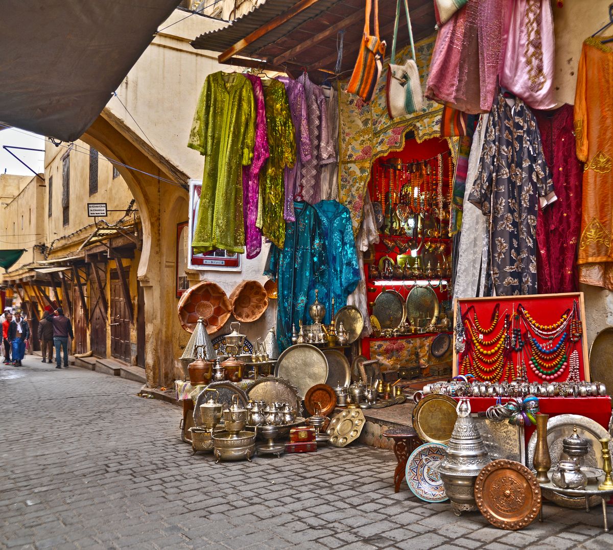 A Fes market stall hung with colorful woven textiles, copper trays and metalware stacked below.