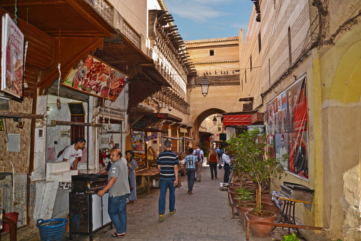 A narrow arched passage through the Fes medina, shopfronts lining the walls and daylight cutting a bright strip down the middle.