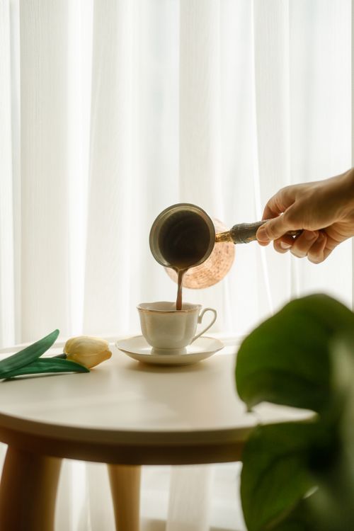 Free Hand pouring hot coffee into a cup with sunlight streaming through curtains, creating a warm ambiance. Stock Photo