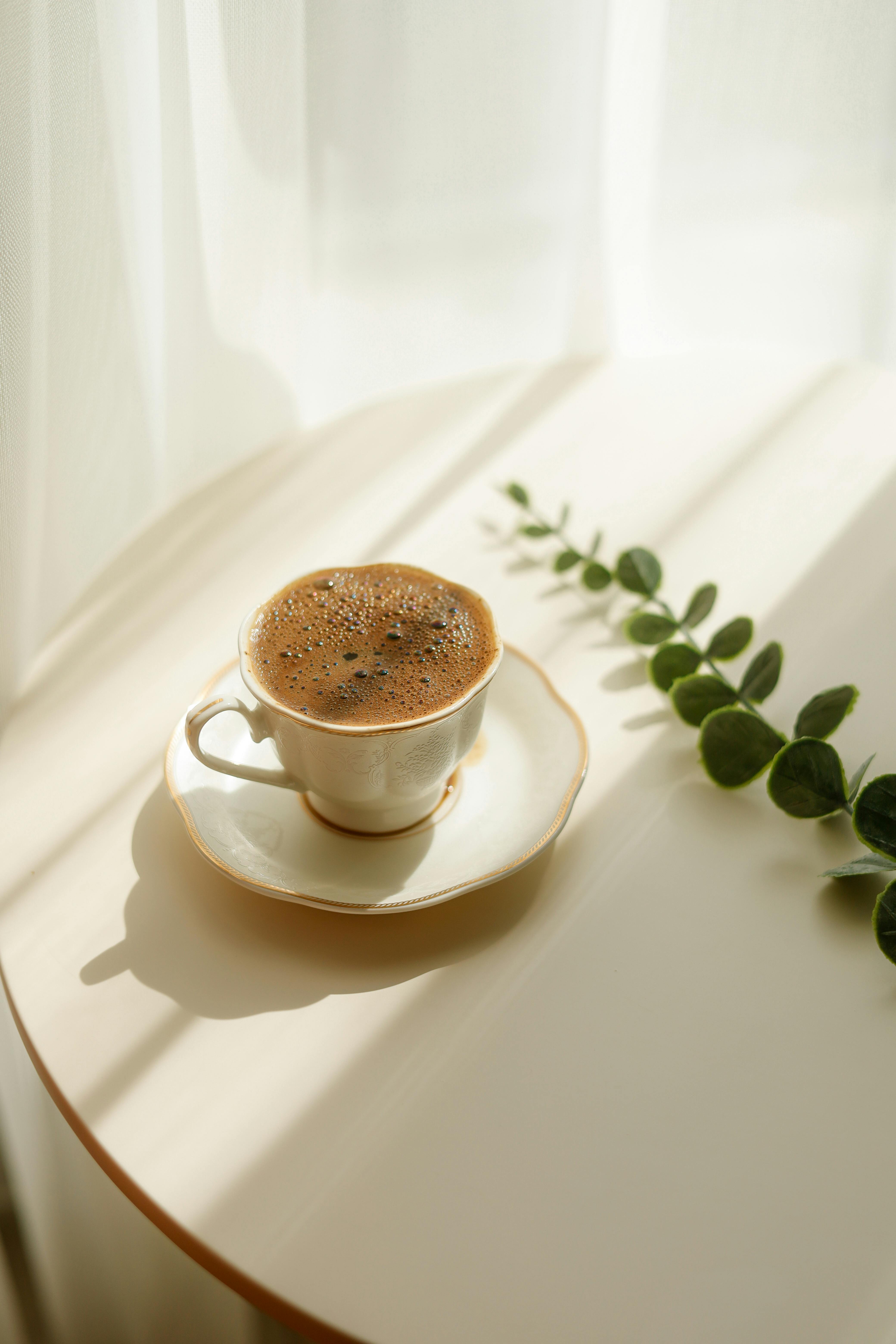 Cup of frothy coffee on table with soft sunlight and green leaf.