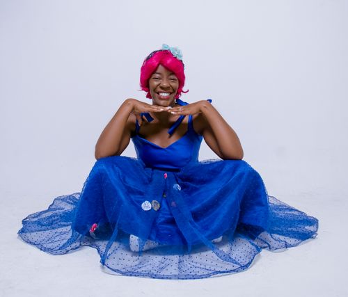 Free Smiling woman in vibrant blue dress with pink wig, sitting indoors. Stock Photo