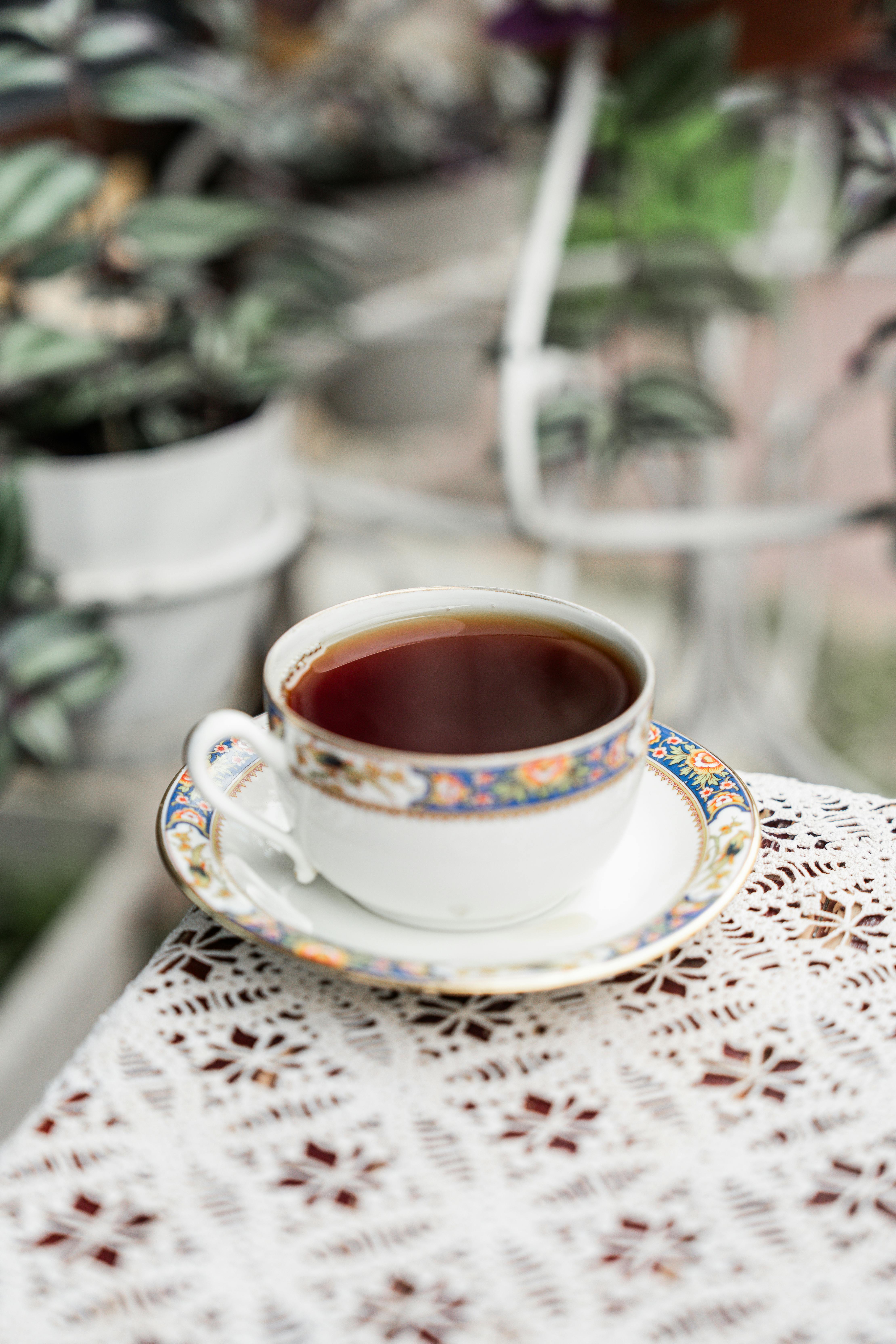 Ornate tea cup on lace tablecloth with blurred plants in background.