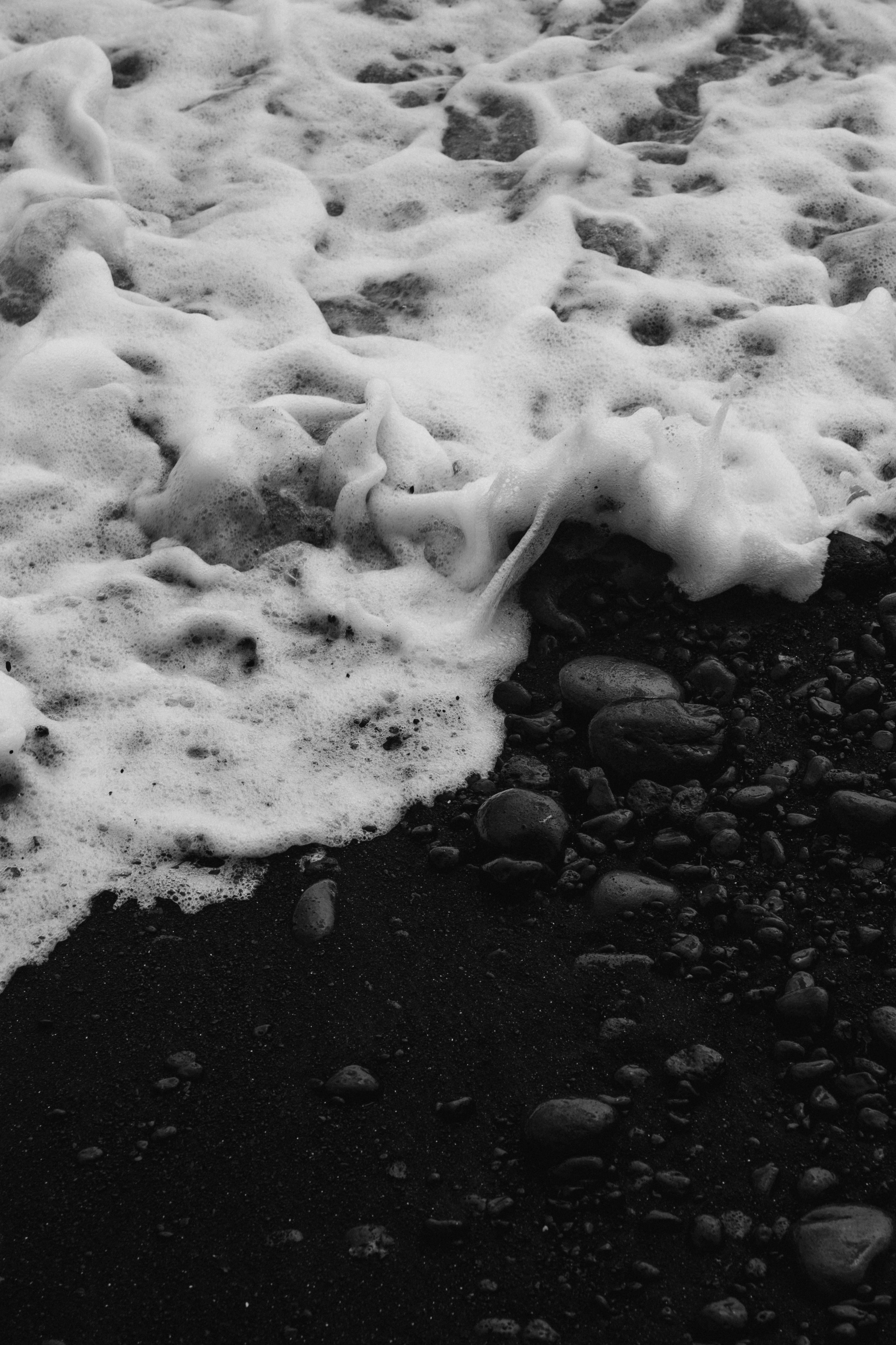 Monochrome image of frothy ocean waves lapping over smooth pebbles on a dark sandy beach.