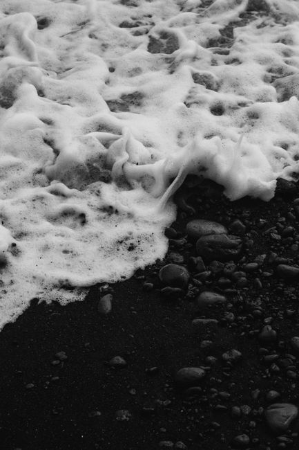 Monochrome image of frothy ocean waves lapping over smooth pebbles on a dark sandy beach.