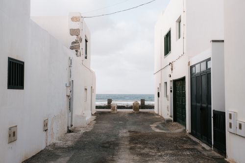 Free A serene coastal alleyway between white buildings leading to the ocean view on a cloudy day. Stock Photo