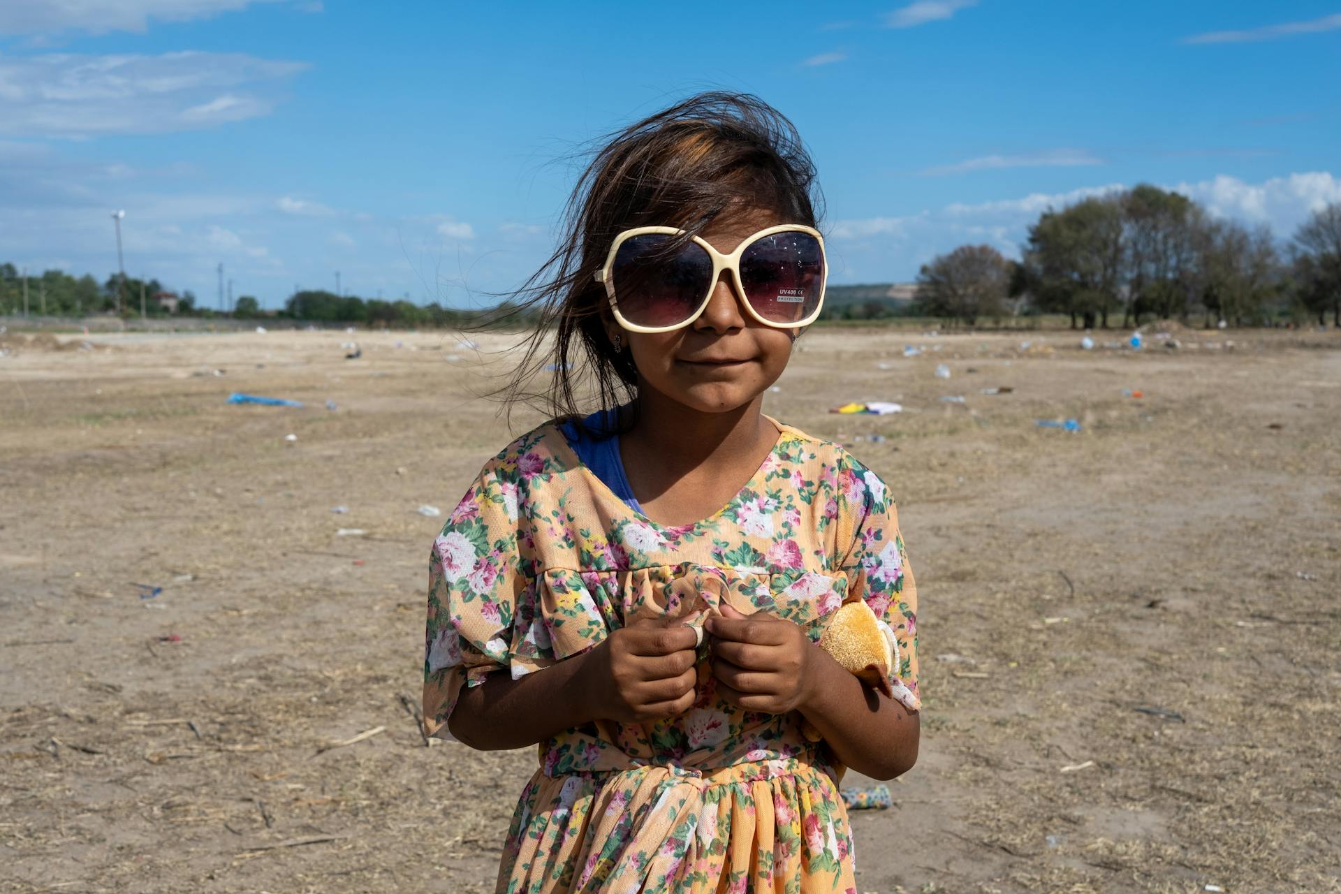 A young girl wearing oversized sunglasses stands confidently in a dry, open field under a clear sky.