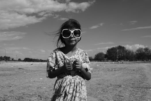 A young girl wearing oversized sunglasses standing on a barren landscape under cloudy skies.
