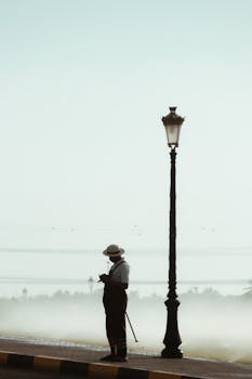 Silhouette of a man with a walking cane beside a street lamp in a foggy landscape at dawn.