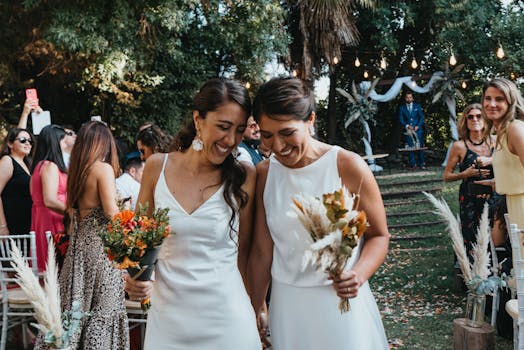 Two brides happily walk down the aisle at an outdoor wedding ceremony surrounded by friends and family.