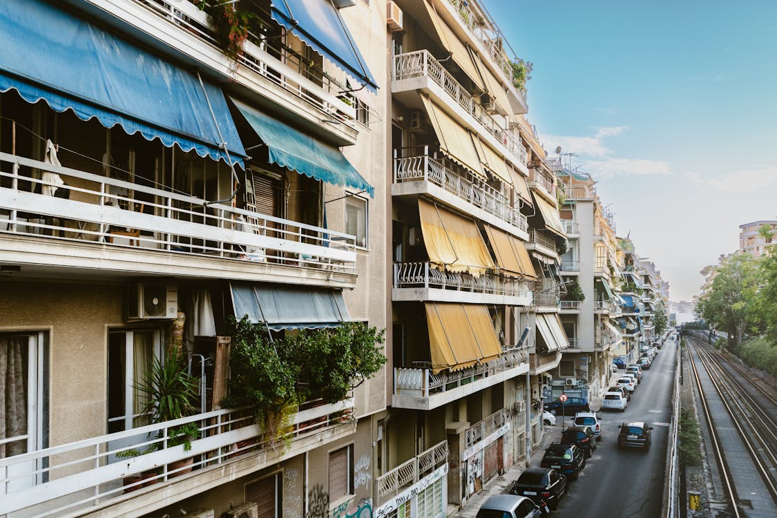Street view of apartment buildings with blue and yellow awnings, parked cars, and a nearby railway track.