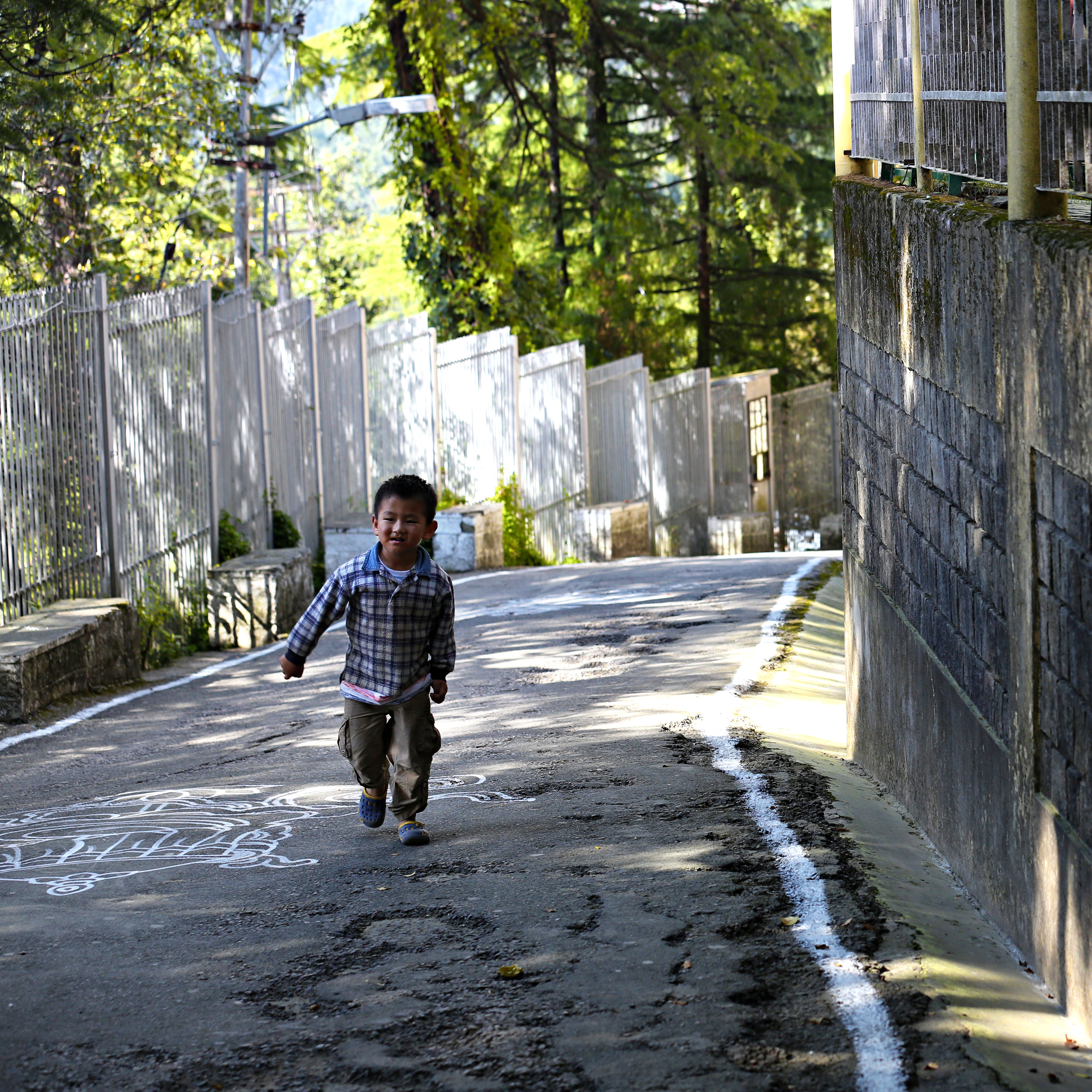 A cheerful boy runs joyfully on a sunlit street surrounded by trees.