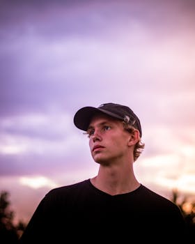 一位戴着帽子的年轻人在日落时分眺望远方，色彩缤纷的天空映衬着他的肖像