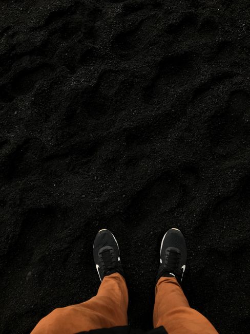 Fotomural Una persona está de pie en una playa de arena negra en Islandia, usando pantalones marrones y zapatillas.