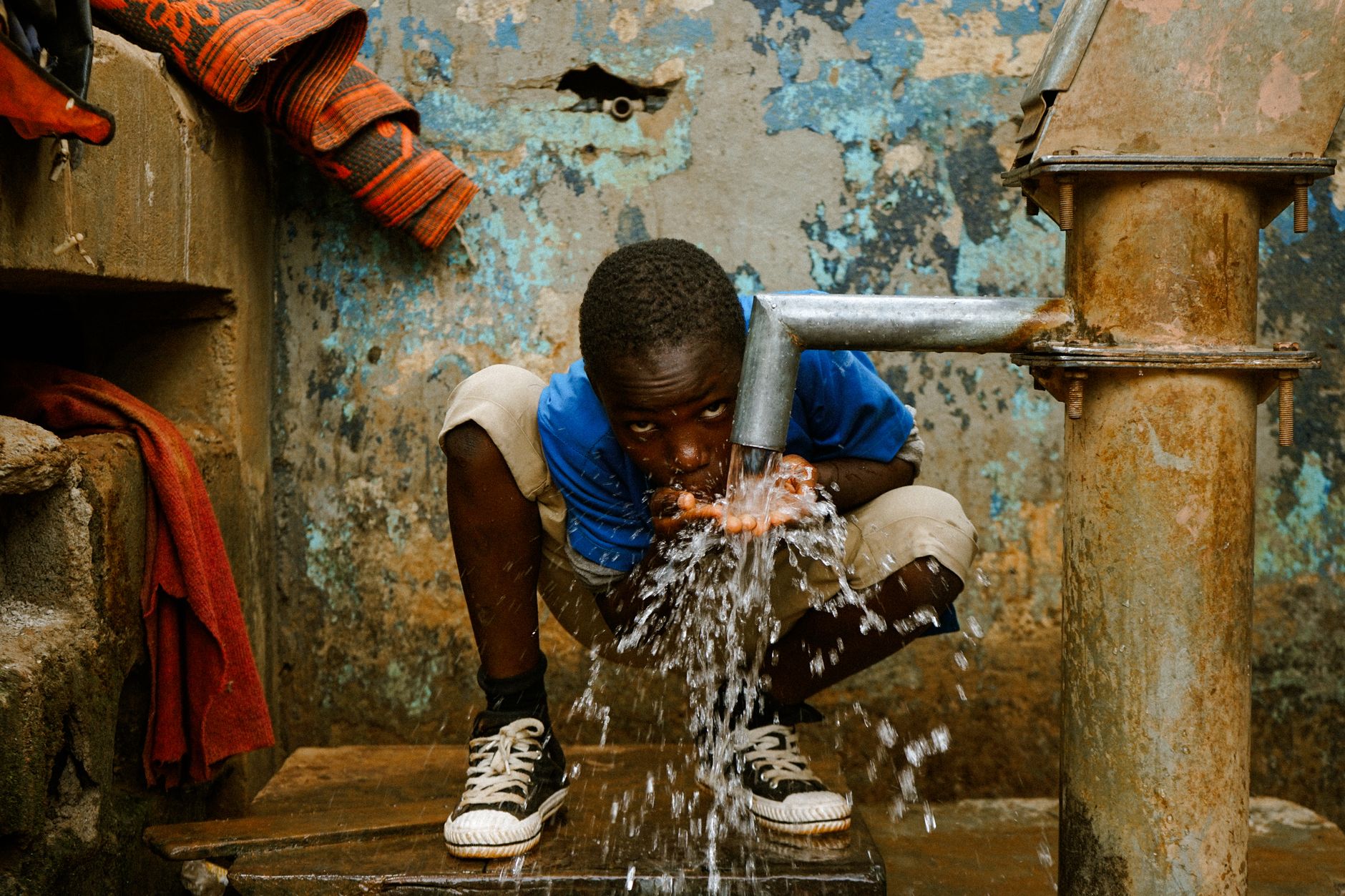 A young boy squats and drinks water from a rusty tap, emphasizing access to clean water.