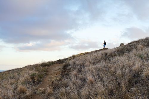 Free A lone hiker stands atop a grassy hill under a vibrant evening sky, capturing a moment of solitude and nature. Stock Photo