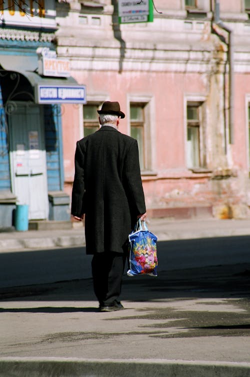 Back view of anonymous elderly man in dark outerwear and hat carrying packet and walking on street against old shabby buildings in city